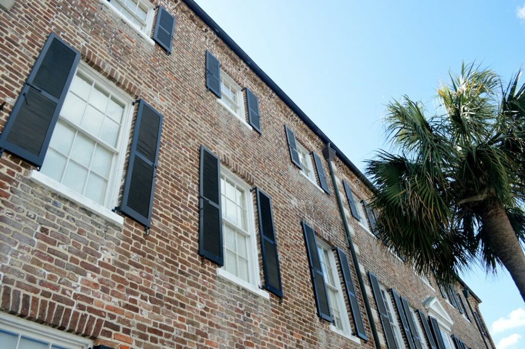 Brick residential building with black shutters beside a palm tree under a clear blue sky