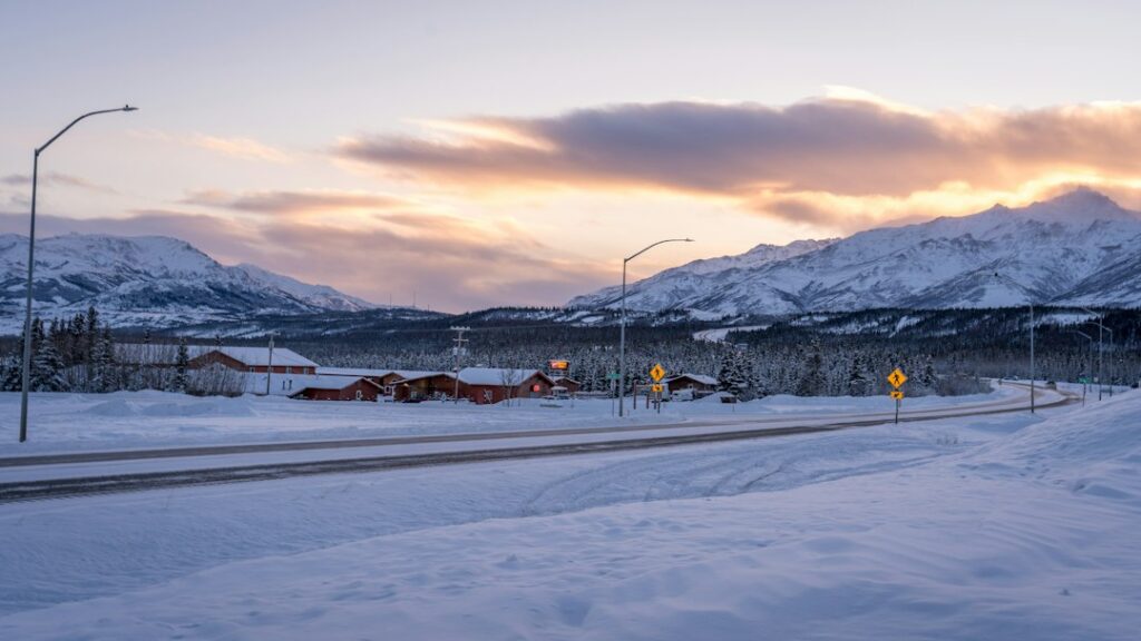 Snowy road leading to buildings in Fairbanks, Alaska, with snow-covered mountains under a sunset sky